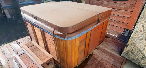 A wooden hot tub with a brown cover, situated on a wooden deck near a rustic wooden wall.